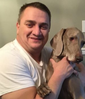 %Focuskw% | Good Tidings Plumbing, Heating &Amp; Cooling A man in a white t-shirt smiles while holding a large gray dog indoors.