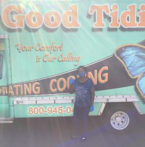 %Focuskw% | Good Tidings Plumbing, Heating &Amp; Cooling Person wearing a face mask and gloves stands in front of a truck with “Good Tidi” and a large blue butterfly graphic on the side in bright sunlight.