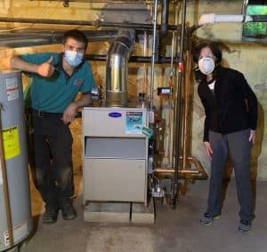 %Focuskw% | Good Tidings Plumbing, Heating &Amp; Cooling Two people wearing masks stand beside a furnace in a basement, with pipes, a water heater, and exposed concrete walls visible.