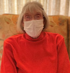 %Focuskw% | Good Tidings Plumbing, Heating &Amp; Cooling An older woman wearing glasses and a white face mask sits indoors on a patterned chair, dressed in a bright red sweater.