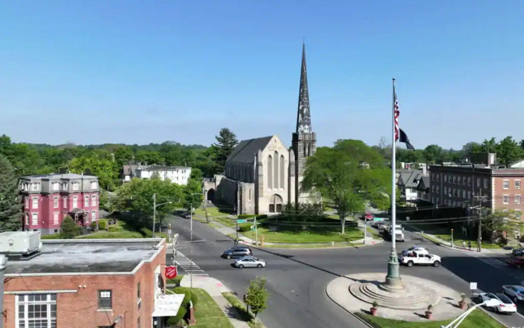 Aerial view of a small town intersection with a church featuring a tall steeple, surrounding buildings, trees, and a flagpole in the center of a roundabout.