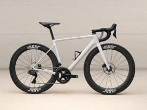 Weißes Rennrad mit hohen Carbon-Laufrädern, auf denen Felgenaufkleber mit der Aufschrift „KEEP GOING“ angebracht sind, vor hellem Hintergrund.