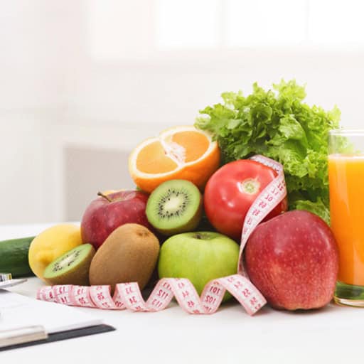 Fresh fruits and vegetables with a measuring tape and glass of orange juice on a white surface.