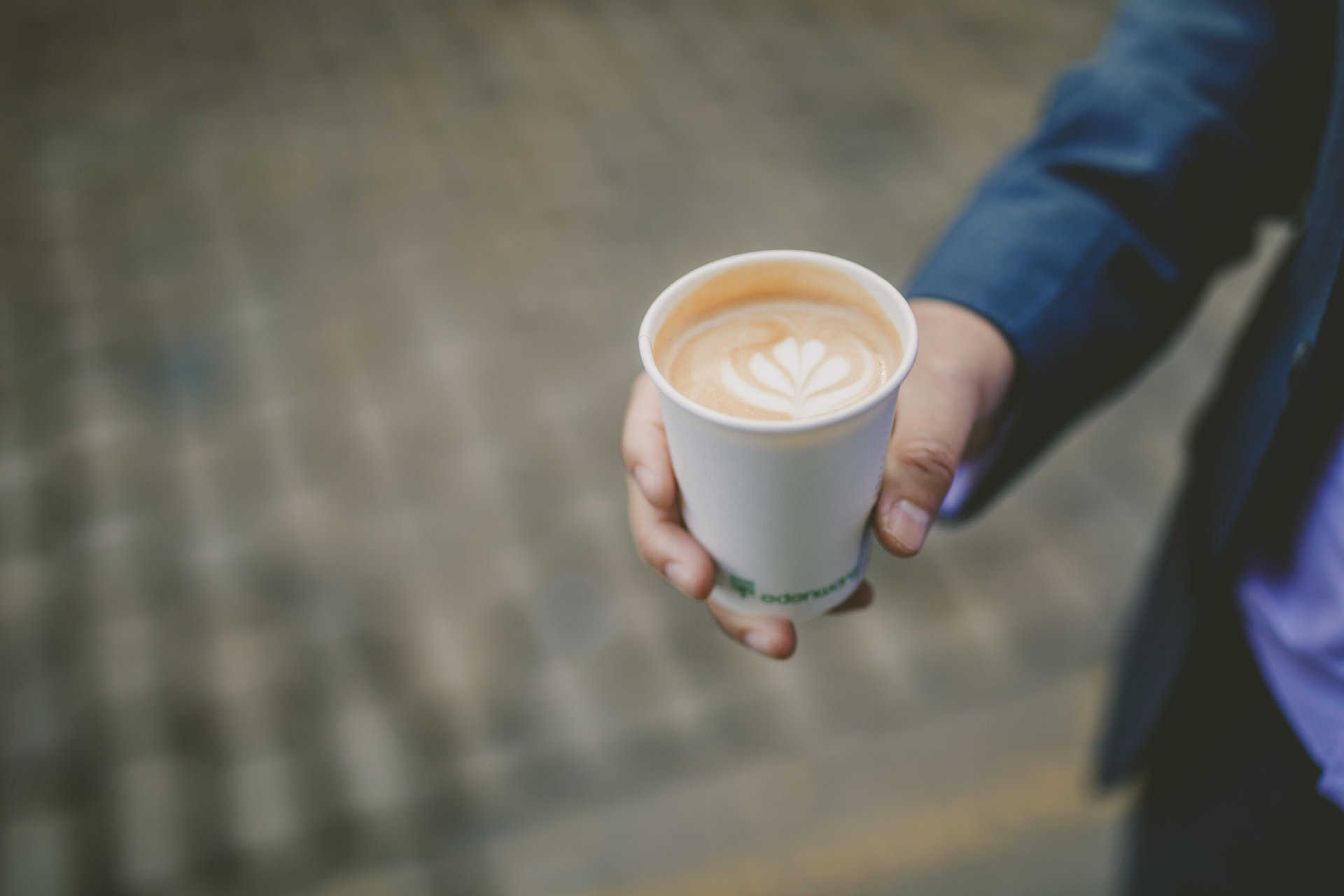 Creamy latte with latte art held by a person in a blue jacket, urban coffee shop setting.