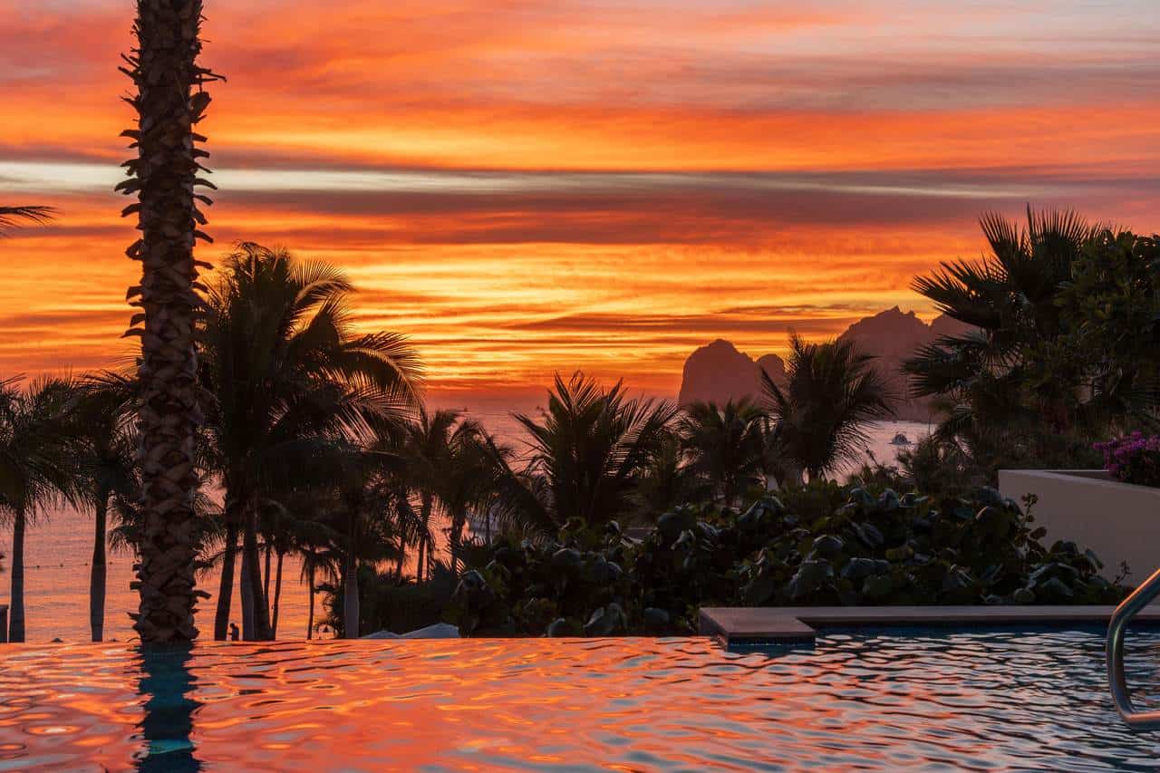 Elegant Sunset View Over A Luxurious Infinity Pool At Hacienda, Featuring Vibrant Orange And Pink Hues Reflecting On The Water With Lush Palm Trees And Scenic Mountains In The Background.
