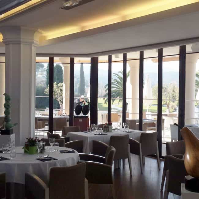 Moderno comedor con grandes ventanales y vistas panorámicas de jardín y palmeras, ideal para reformas integrales en hostelería.