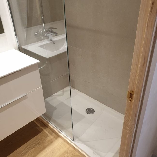 Reforma de ducha moderna con mampara de vidrio y azulejos en tonos beige.