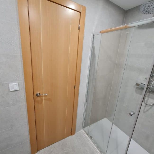 Puerta de madera en baño con ducha y azulejos grises, reforma de baños por Hammerman Reformas.