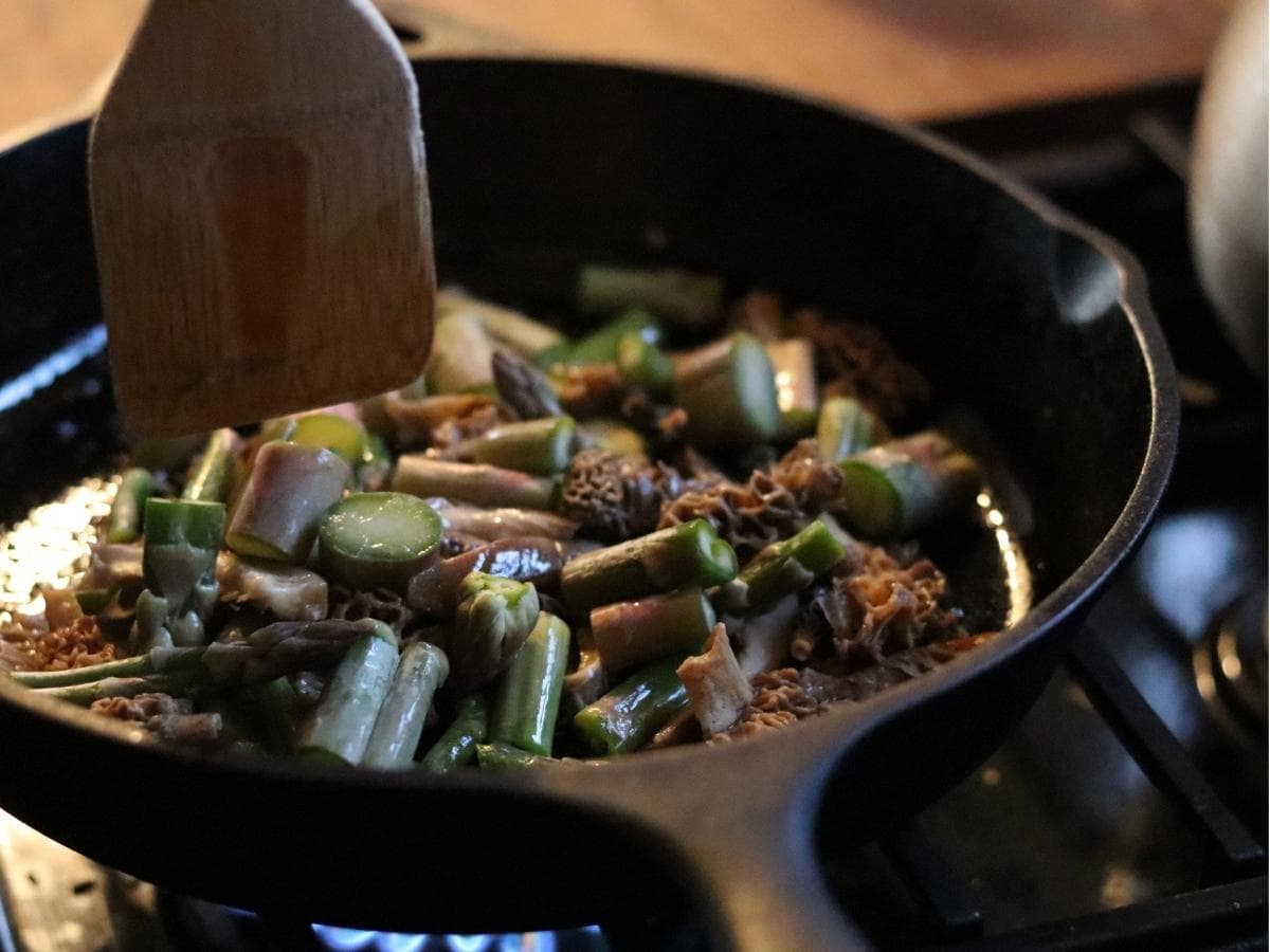 sautéed morels and asparagus cooking in a cast iron pan with a wooden spatula and blue flame underneath