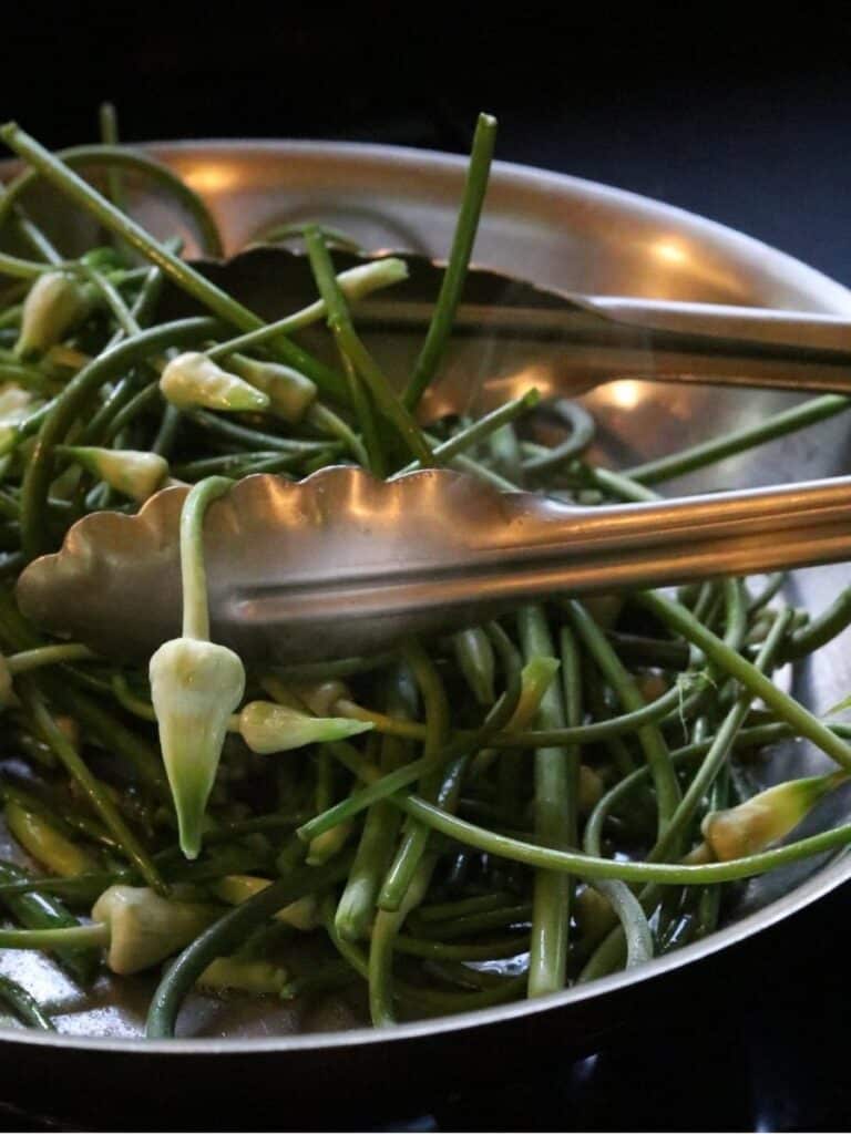 Turning sautéed garlic scapes in pan with tongs