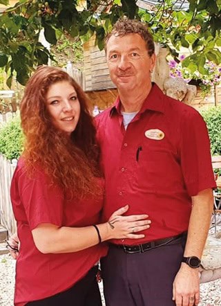 Corinna-Rainer_320x440 Eine Frau mit langen, roten Locken und ein Mann mit kurzen Locken, beide in roten Hemden, stehen zusammen im Freien. Die Frau lächelt und hat ihren Arm um den Mann gelegt. Sie befinden sich in einer Gartenanlage mit viel Grün und Holzkonstruktionen.
