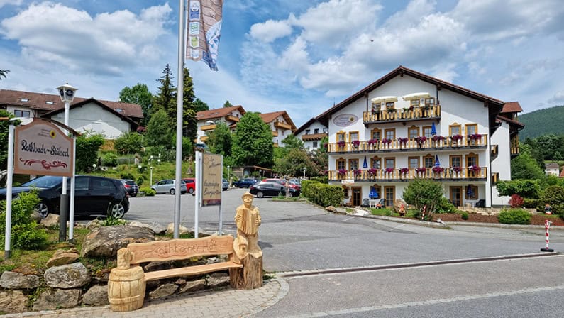 Einfahrt-neu_795x448 Ein uriges Gasthaus im bayerischen Stil mit weißer Fassade und Blumenkästen auf jedem Balkon. Im Vordergrund, neben dem Gasthausschild, steht eine Holzbank mit einer geschnitzten Figur. Die Szene spielt an einem leicht bewölkten Tag.