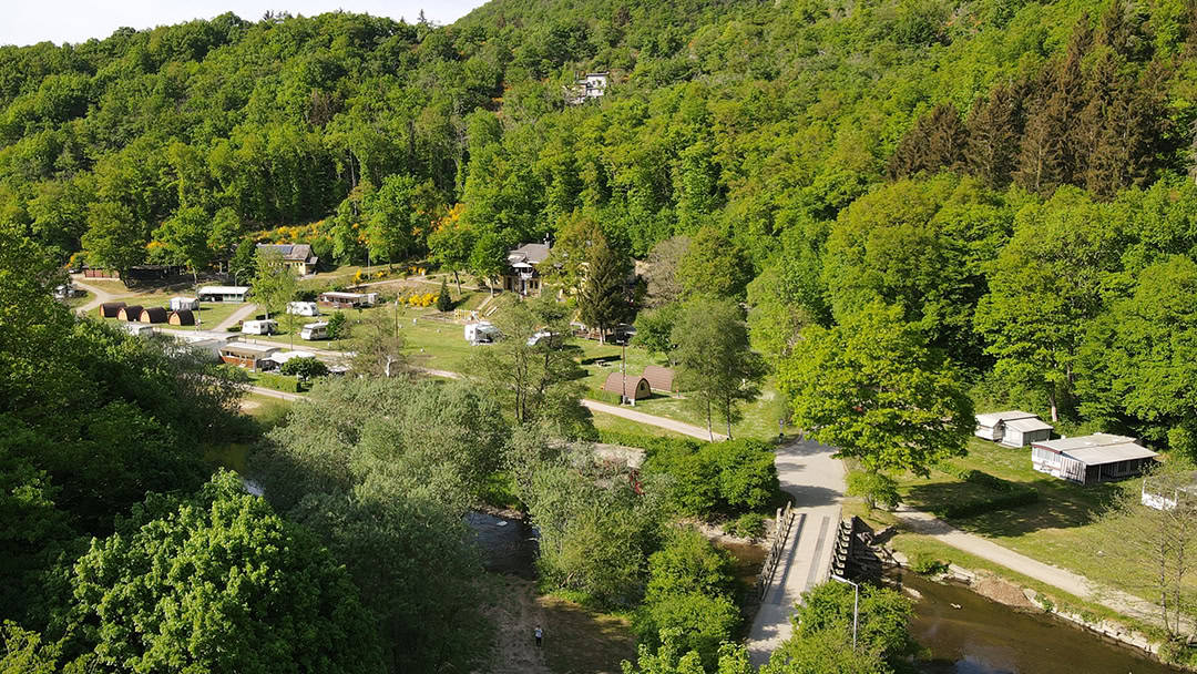 Kauterbach_1080608 Luftaufnahme eines üppigen, grünen Waldes mit verstreuten Häusern und Wohnmobilen. Eine kleine Straße führt über eine Brücke über einen Fluss, umgeben von dichten Bäumen. Das leuchtende Grün vermittelt an einem klaren Tag den Eindruck einer friedlichen, ländlichen Umgebung.