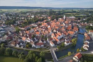 Luftbild_Riedlingen - Heimat erleben mit Magazinen und Katalogen als ePaper Luftaufnahme einer malerischen Stadt mit kreisförmig angeordneten Gebäuden mit roten Dächern, umgeben von üppigem Grün und einem gewundenen Fluss. In der Mitte steht ein markanter Kirchturm, und eine Brücke überquert den Fluss.