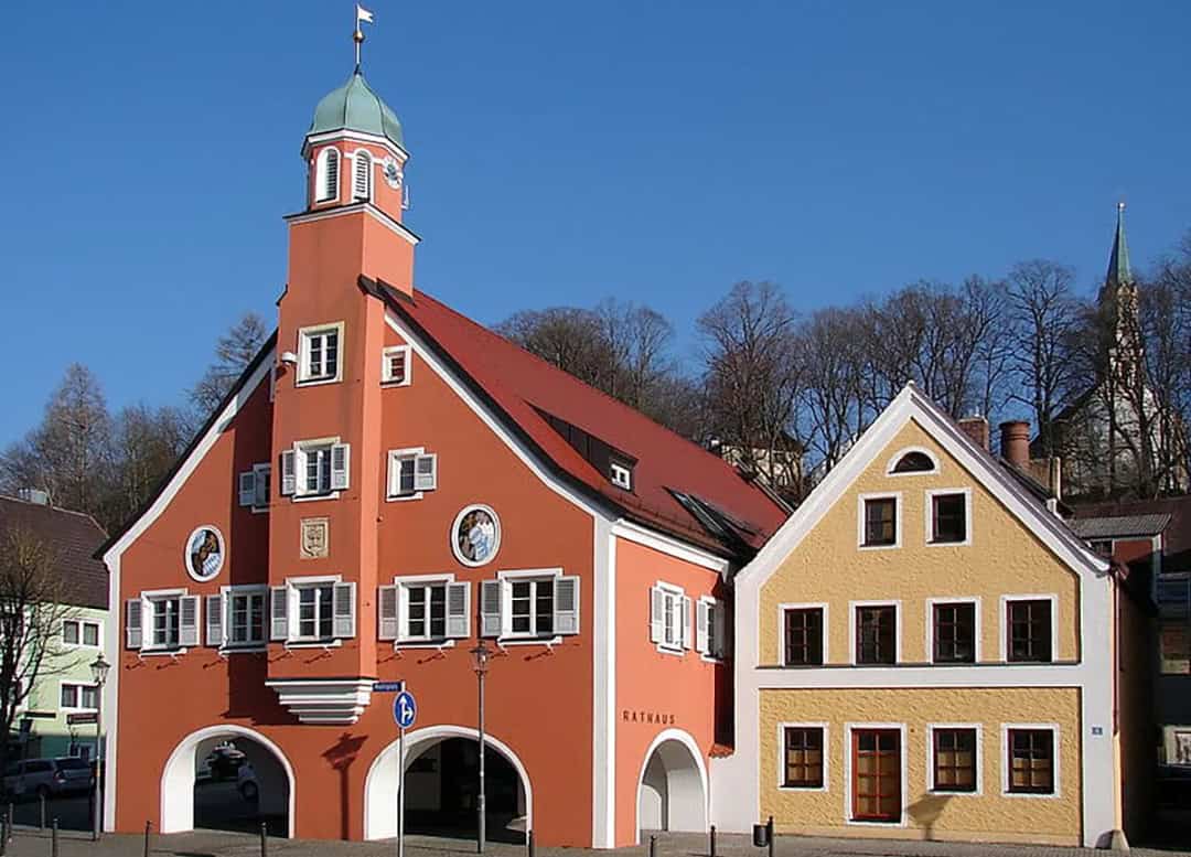 Mainburg_Rathaus_1080x Ein malerisches europäisches Gebäude mit roter Fassade, Glockenturm und gewölbtem Eingang steht vor einem klaren blauen Himmel. Daneben steht ein gelbes Gebäude mit Fensterläden. Im Hintergrund sind Bäume und ein Kirchturm zu sehen.