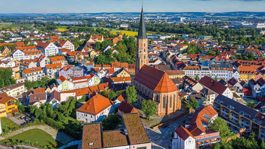 Dingolfing_1080 Luftaufnahme einer malerischen europäischen Stadt mit farbenfrohen Häusern und einer großen Kirche mit hohem Turm. Die Stadt ist von grünen Feldern umgeben und in der Ferne ist unter einem teilweise bewölkten Himmel ein Fluss zu sehen.