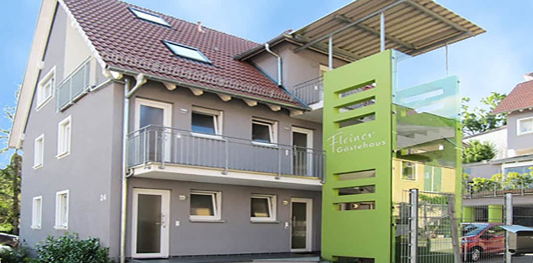 Gaestehaus_Fleiner_Ansicht_1080 Ein modernes, dreistöckiges Gästehaus mit beigefarbener Fassade und Ziegeldach. Das Gebäude verfügt über Balkone und einen grünen Aufzugsschacht mit Glaspaneelen. Der Himmel ist klar und blau.