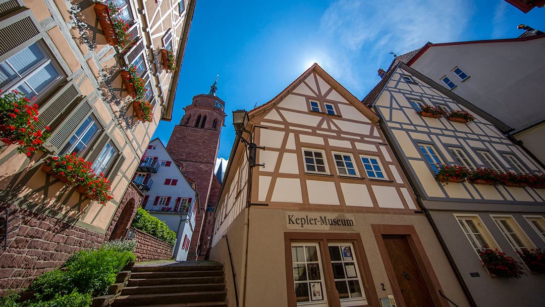 Weil-der-Stadt_Achim_Mende_2_1080 Eine sonnenbeschienene Straße in Weil der Stadt, Deutschland, mit blumengeschmückten Fachwerkhäusern. Das Kepler Museum ist zu sehen, im Hintergrund ein Kirchturm unter einem strahlend blauen Himmel.