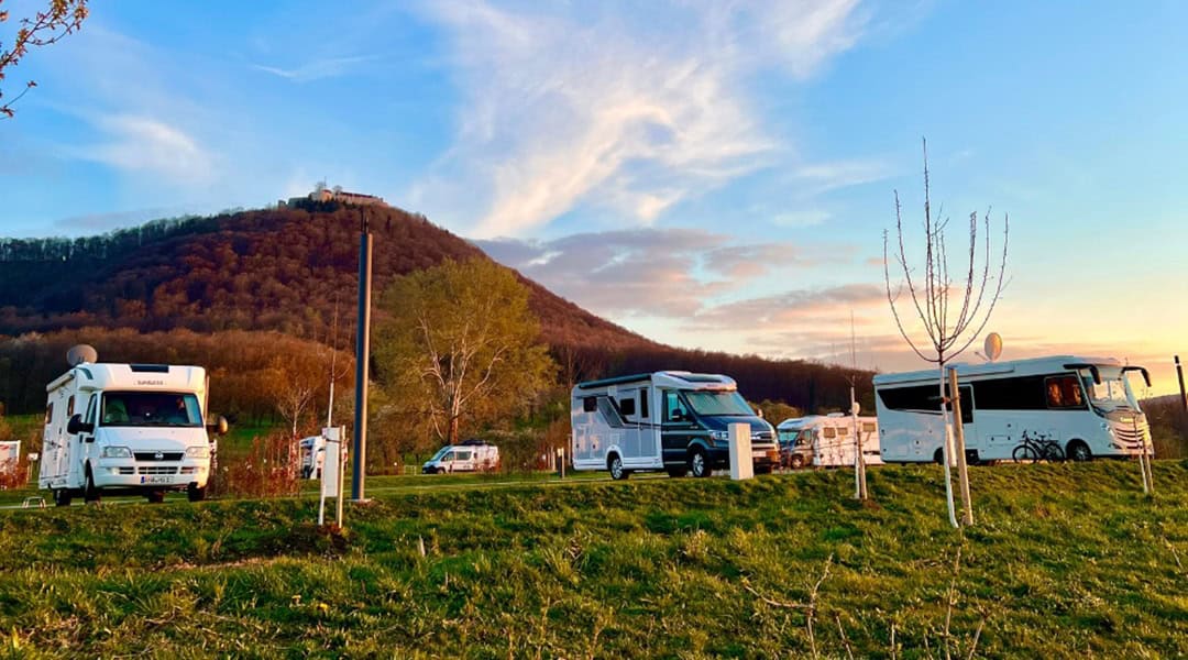 Abendstimmung-Womo-PLatz_Panorama-Therme_Linha_Reeth Drei Wohnmobile parken bei Sonnenuntergang auf einer Wiese. Im Hintergrund sind ein Hügel und vereinzelte Bäume zu sehen. Der Himmel ist teilweise bewölkt und strahlt warm, was eine ruhige Atmosphäre schafft.