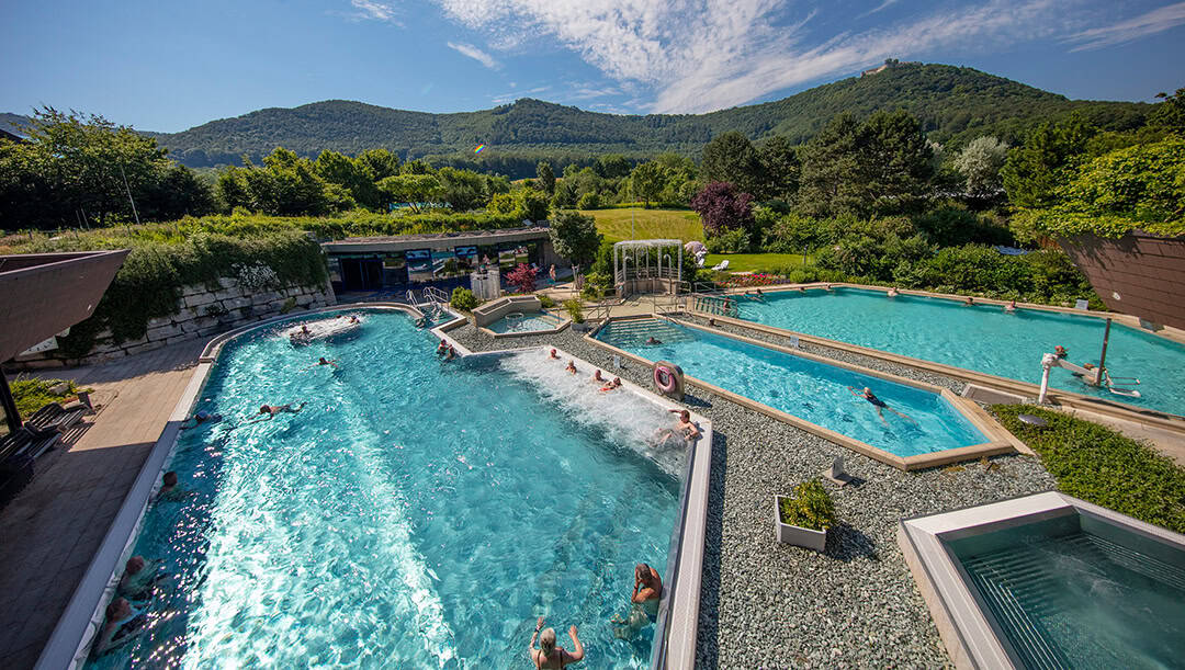 Beuren_Mende_1080 Außenpools mit Menschen, die das Wasser genießen, umgeben von üppigem Grün und Hügeln unter blauem Himmel. Der Poolbereich verfügt über verschiedene Bereiche mit türkisfarbenem Wasser und angelegten Rändern.