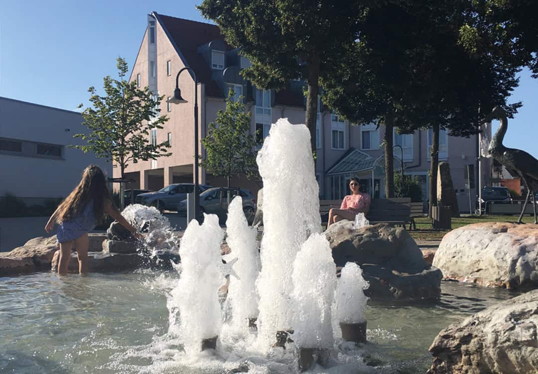 PHS_Hotel-mit-Brunnen_1080 Ein junges Mädchen spielt an einem sprudelnden Brunnen in einem sonnendurchfluteten Park. Im Hintergrund sitzt eine Person auf einer Bank in der Nähe von Bäumen. In der Nähe steht eine Storchenskulptur. Unter einem strahlend blauen Himmel ist eine Reihe von Gebäuden zu sehen.