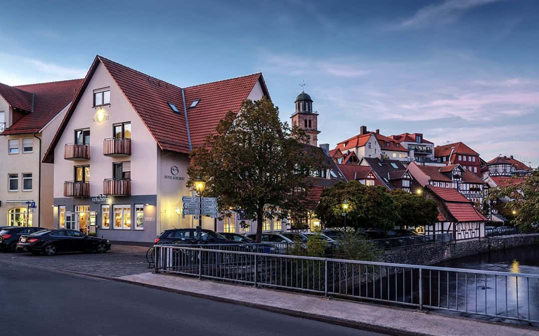 Romantik-Hotel-Schubert-in-Lauterbach-1080 Dämmerungsansicht eines europäischen Dorfes mit traditionellen Häusern und schrägen roten Dächern. Ein kleiner Kanal verläuft entlang der Straße, schwach beleuchtete Lampen erhellen die Gegend. Im Hintergrund steht ein hoher Kirchturm.