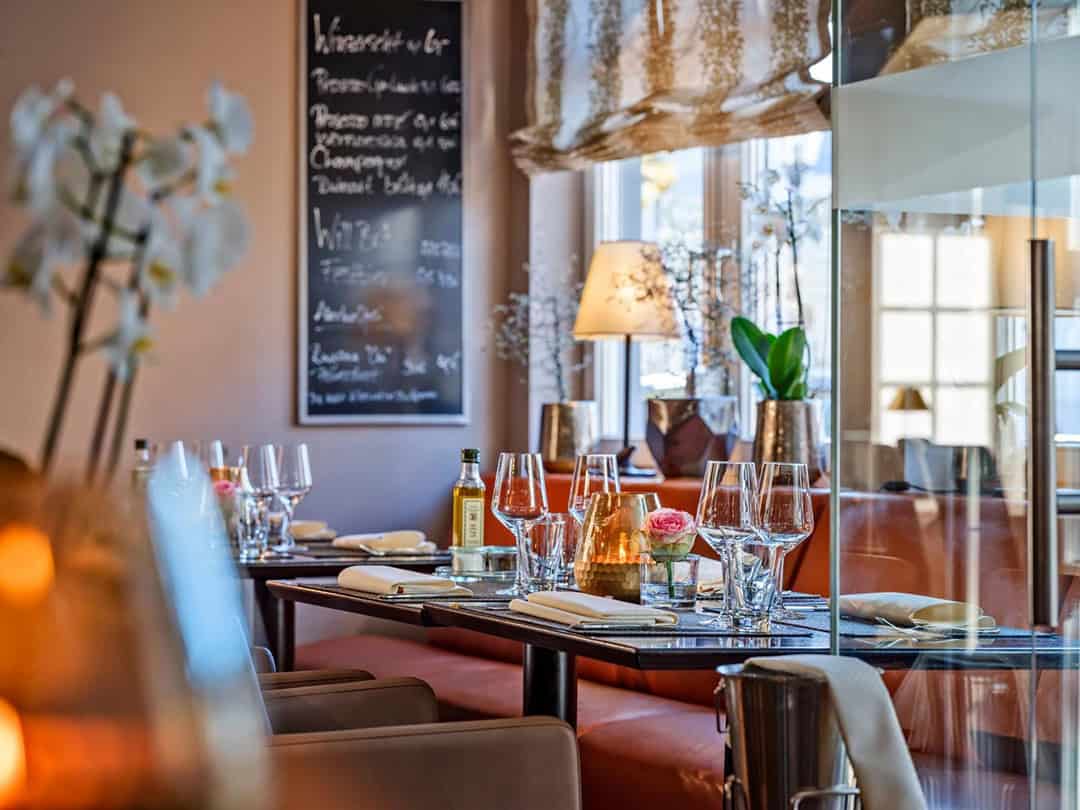 Romantik-Hotel-Schubert-in-Lauterbach-Restaurant-Schuberts-1080 Ein gemütliches Restaurant mit gedecktem Tisch, Weingläsern, Servietten und Blumenschmuck. Im Hintergrund ist eine Speisekarte auf Kreidetafel zu sehen. Warme Beleuchtung und geschmackvolle Dekoration schaffen eine einladende Atmosphäre.