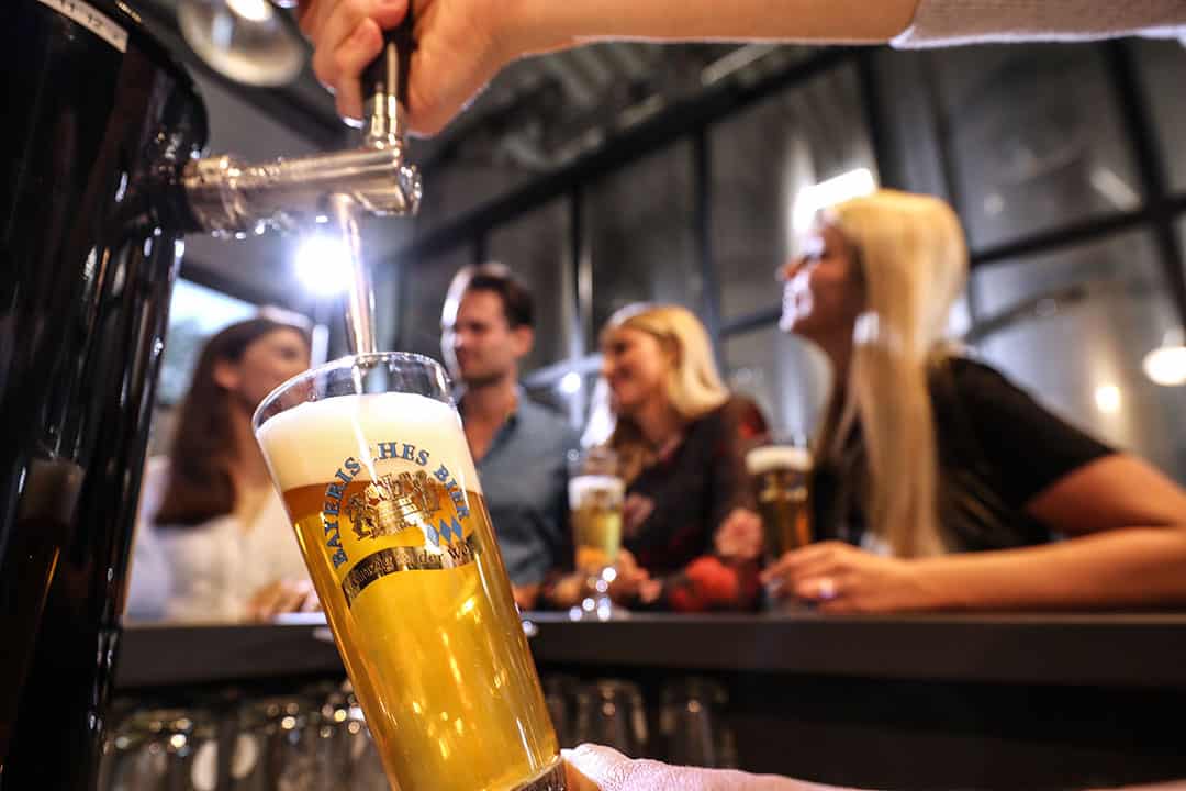 bayerisches_bier_tresen_zapfhahn_1080 Eine Hand schenkt ein Glas Bier aus einem Zapfhahn ein, während im Hintergrund Menschen an einer Bar Getränke genießen und sich unterhalten. Die Szene ist lebhaft und gesellig, und der Fokus liegt auf dem frisch ausgeschenkten Bier.