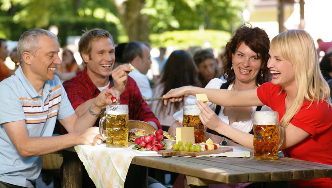 bg06_brotzeit05_1080 Eine Gruppe von vier Personen sitzt im Freien an einem hölzernen Picknicktisch und genießt Krüge Bier und eine Platte mit Snacks, darunter Käse, Radieschen und Trauben. Sie lächeln und scheinen sich angeregt zu unterhalten. Im Hintergrund sind Bäume und Menschen zu sehen.