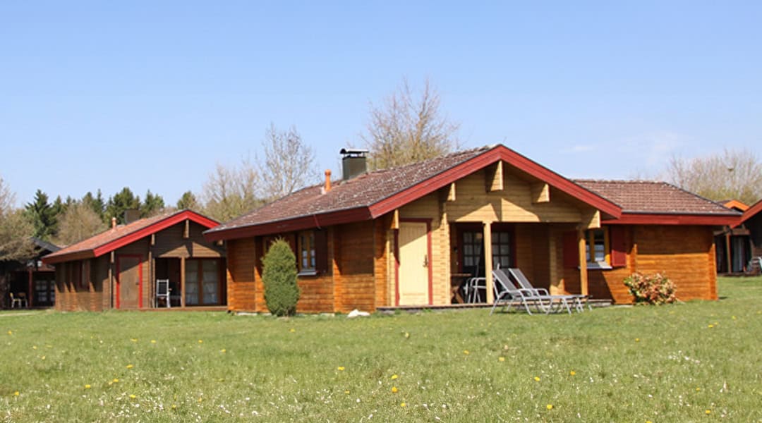 Lauterdoerfle_Header_1080 Eine Holzhütte mit rotem Dach und Veranda steht auf einer Wiese unter strahlend blauem Himmel. Zwei Liegestühle stehen in der Nähe des Eingangs, im Hintergrund ist eine weitere ähnliche Hütte zu sehen. Bäume und Sträucher umgeben das Gelände.