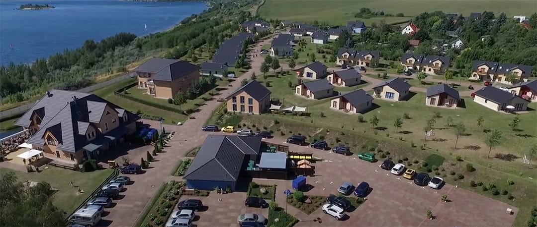 Seepark-Luftbild-1080 Luftaufnahme eines Seebads mit mehreren modernen Häusern und Gebäuden, umgeben von Grün. Autos parken entlang der gepflasterten Wege, und auf der linken Seite ist das blaue Wasser des Sees sichtbar, gesäumt von Bäumen.