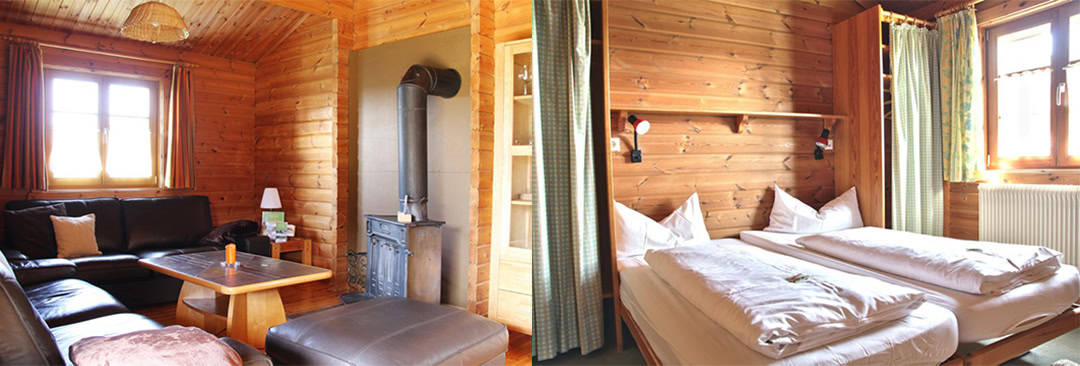Zimmer_1080 Das Innere einer gemütlichen Hütte: links ein Wohnbereich mit schwarzem Ledersofa, Holztisch und Metallofen vor holzgetäfelten Wänden; rechts ein Schlafzimmer mit zwei Einzelbetten, Kissen, Holzwänden und Fenstern mit Vorhängen.