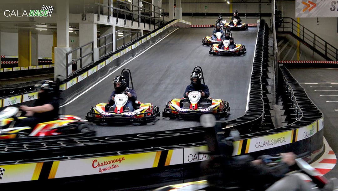 CalaRace_Bahn1_1080 Menschen fahren Go-Karts auf einer Indoor-Bahn mit mehreren Spuren. Die Strecke ist ansteigend und von Barrieren umgeben. Die Fahrer tragen Helme und fahren in rot-gelben Karts. Im Hintergrund sind Beschilderungen und Sicherheitsmarkierungen zu sehen.