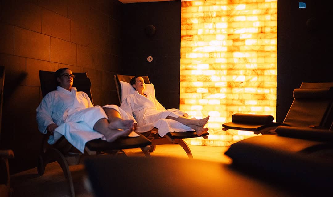 CalaSpa02_1080 Zwei Menschen in weißen Bademänteln entspannen auf Liegestühlen in einem schwach beleuchteten Spa-Raum. Eine warme, leuchtende Salzwand sorgt für stimmungsvolles Licht und schafft eine ruhige Atmosphäre.