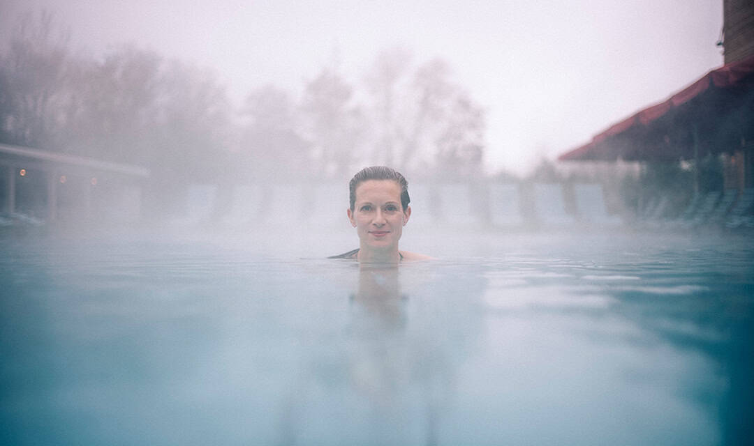 CalaSpa03_1080 Eine Person schwimmt in einem Außenpool, umgeben von Nebel. Ihr Kopf ragt über Wasser, im Hintergrund sind Liegestühle zu sehen. Die Atmosphäre ist heiter und neblig und schafft eine ruhige Atmosphäre.