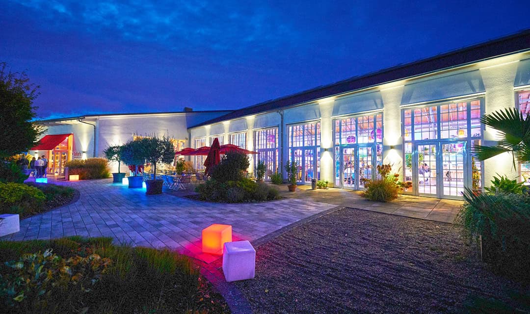 Orangerie01_1080 Abendszene im Freien: Ein modernes Gebäude mit großen, weiß beleuchteten Glasfenstern. Der Weg ist mit bunten Würfellichtern in Rot, Blau und Grün beleuchtet. Bäume und Tische mit roten Sonnenschirmen sind zu sehen. Der Himmel ist tiefblau.
