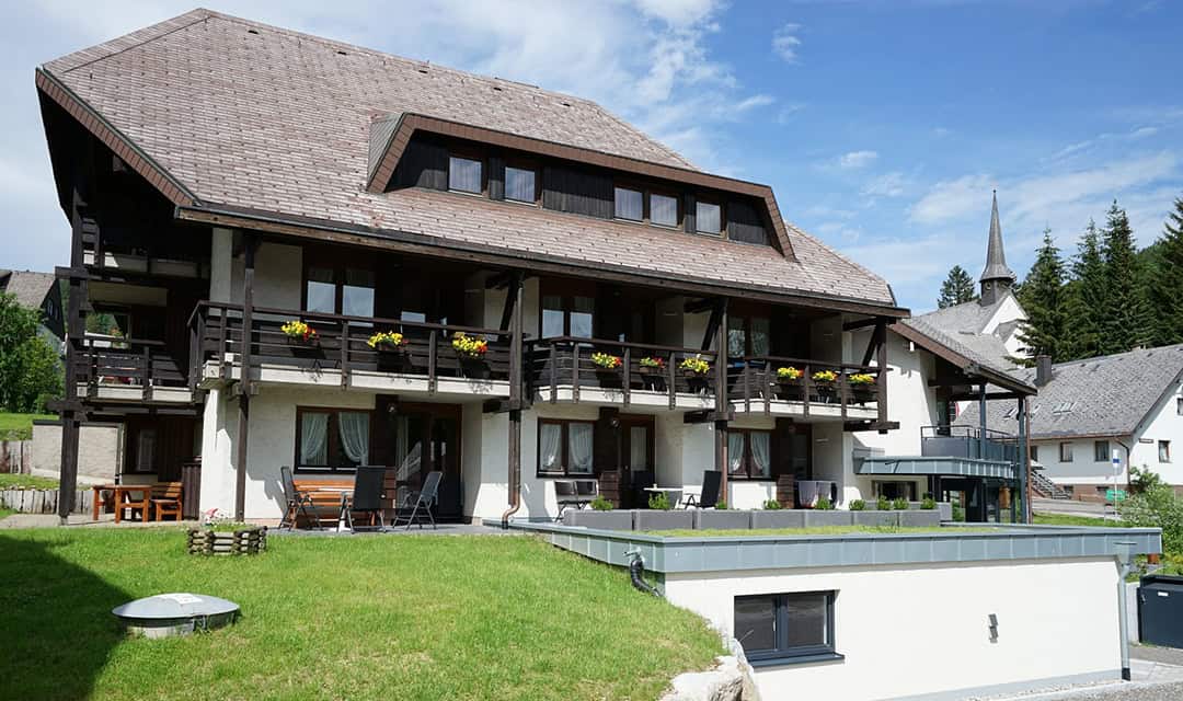 Schwarzwaldmaedel_Aussen_1080 Ein großes Haus im Chalet-Stil mit steilem Dach und Holzbalkonen, umgeben von grünem Gras. Auf den Balkonen blühen gelbe Blumen, im Hintergrund ist ein Kirchturm zu sehen. Der Himmel ist teilweise bewölkt.