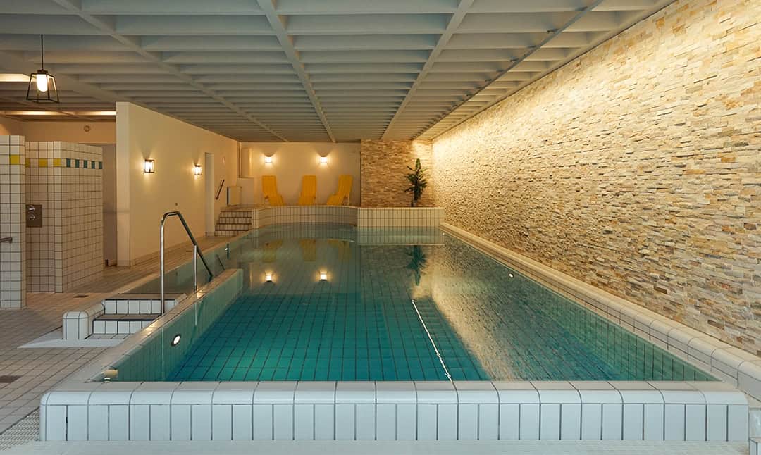 Schwarzwaldmaedel_Schwimm_1080 Innenpool mit blauen Fliesen, umgeben von beigen Steinmauern und einer weiß gefliesten Terrasse. In der Ecke stehen mehrere gelbe Liegestühle. Die Decke ist mit einem Gittermuster versehen, und der Raum wird durch Wandleuchten sanft beleuchtet.