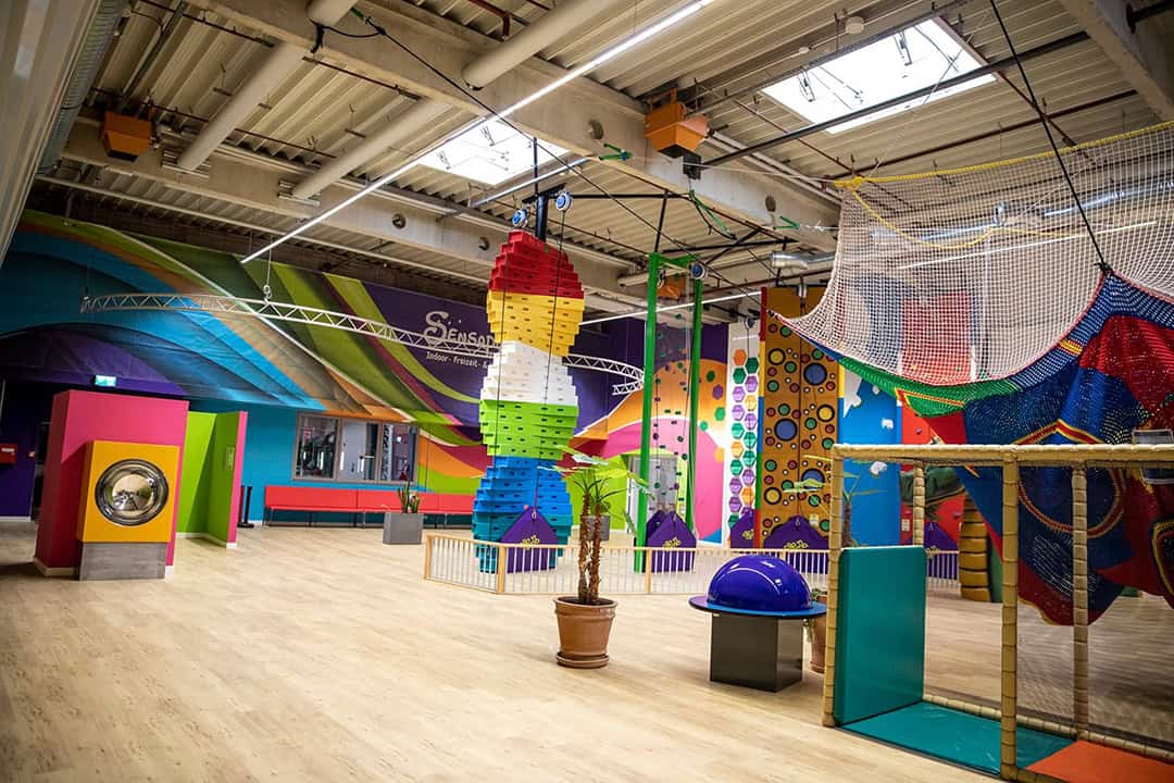 Sensapolis_1080 Farbenfroher Indoor-Spielplatz mit Klettergerüsten, Hängenetzen und farbenfrohen Wandmalereien. Der Bereich umfasst eine skurrile, bunte Raketenskulptur und verschiedene interaktive Elemente für Kinder – alles unter Oberlichtern und freiliegenden Decken.