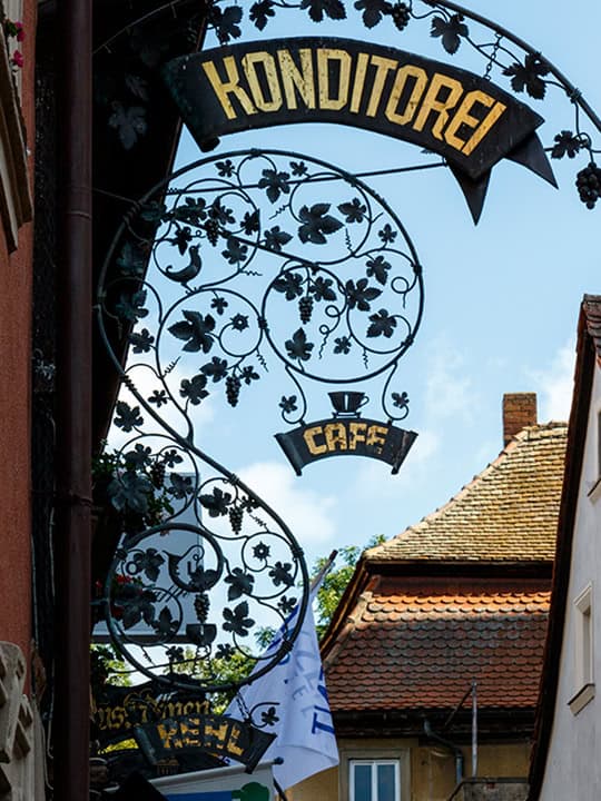 Cafe03_540 Auf einem kunstvollen Schild aus Schmiedeeisen mit floralen Verzierungen steht in goldenen Buchstaben „Konditorei Café“ vor blauem Himmel. Im Hintergrund sind Dächer und Flaggen zu sehen.