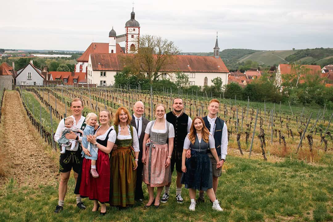 Familie-II_1080 Eine Gruppe von neun Personen steht in einem üppigen Weinberg. Sie tragen traditionelle bayerische Kleidung. Im Hintergrund ist ein Dorf mit rot gedeckten Häusern und einer Kirche zu sehen. Der Himmel ist bedeckt.