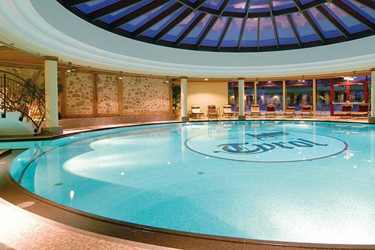 Innenpool_540 Der kreisförmige Innenpool ist von Steinmauern und einer Glaskuppel umgeben. Liegestühle säumen den Pool, und in der Mitte des Wassers befindet sich ein Logo. Sanfte Beleuchtung sorgt für eine ruhige Atmosphäre.