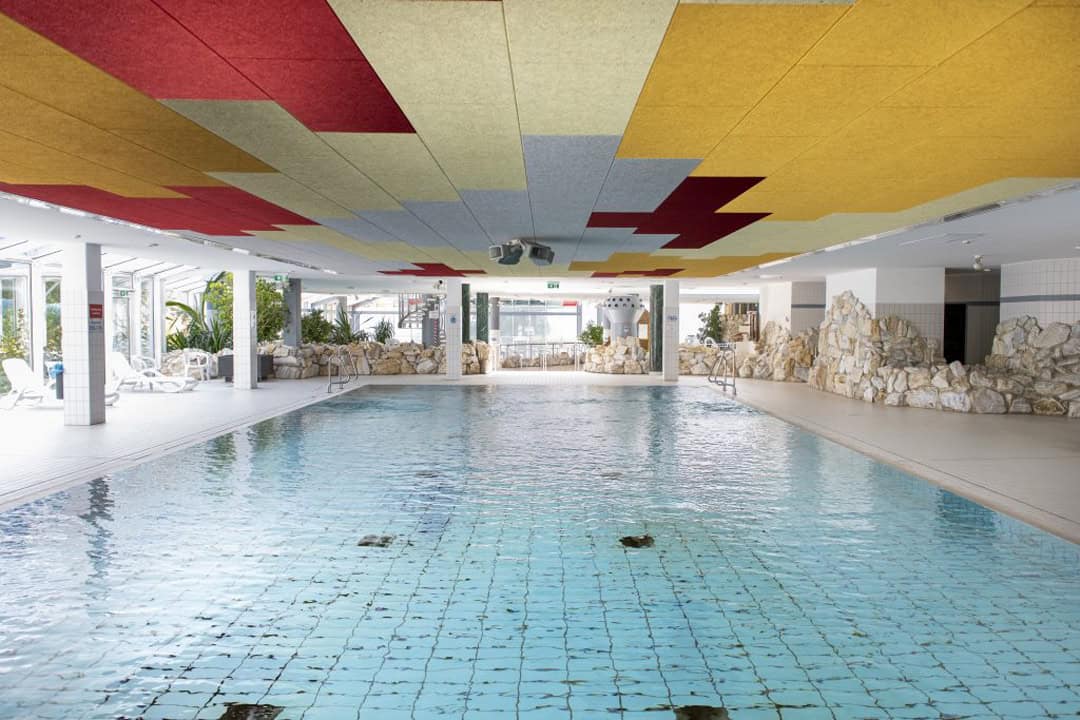 Schwimmbad-Tholey-11-1080 Innenpool mit gefliestem Boden, umgeben von weißen Wänden und dekorativen Felselementen. Die Decke ist mit farbenfrohen Akustikplatten in Gelb, Rot und Grau ausgestattet. Liegestühle und Topfpflanzen säumen den Poolbereich an den großen Fenstern.
