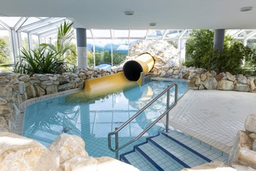 Schwimmbad-Tholey-13-360 Innenpoolbereich mit einer gelben Rutsche, die aus einer Felsstruktur hervorragt. Der Pool ist von Felsen und Grün umgeben, und blau geflieste Stufen führen ins Wasser. Große Fenster bieten Ausblicke auf die Landschaft.