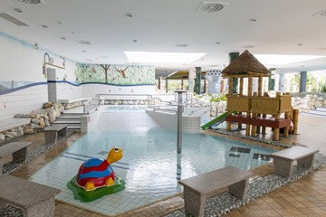 Schwimmbad-Tholey-15-360 Ein überdachter Kinderpoolbereich mit einer bunten Schildkröte. Es gibt ein flaches Becken mit einem kleinen Holzspielgerät mit Rutsche. Wandmalereien zeigen Landschaften. Sitzbänke umgeben den Pool.