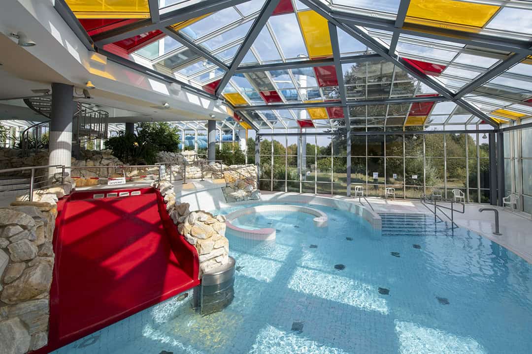 Schwimmbad-Tholey-16-1080 Hallenbad mit klarem Glasdach und farbenfrohen roten und gelben Paneelen. Es verfügt über eine rote Rutsche, Treppen und Sitzbereiche, umgeben von üppigem Grün und großen Fenstern mit Blick auf die Bäume im Freien. Steinakzente schmücken den Poolbereich.