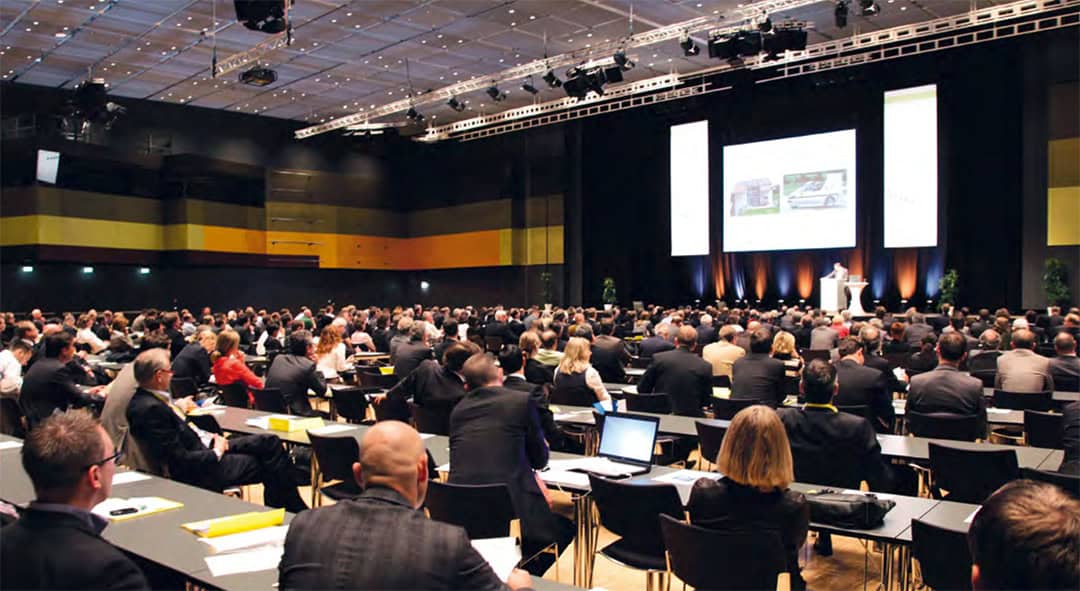 Eine große Konferenz mit zahlreichen Teilnehmern, die an Tischen in einem geräumigen Saal sitzen. Ein Redner präsentiert auf der Bühne, hinter ihm läuft eine Leinwand. Das Publikum, in Business-Kleidung gekleidet, verfolgt die Präsentation aufmerksam.