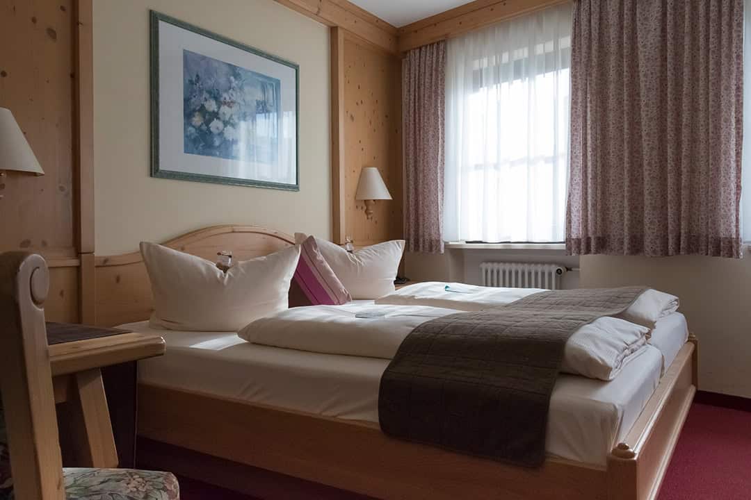 Zimmer07_1080 Ein gemütliches Hotelzimmer mit einem Doppelbett mit weißer Bettwäsche und braunen Decken. Das Zimmer verfügt über Holzwände, ein gerahmtes Blumengemälde und ein Fenster mit hellen Vorhängen, das Tageslicht hereinlässt. Neben dem Bett steht ein Holzstuhl.