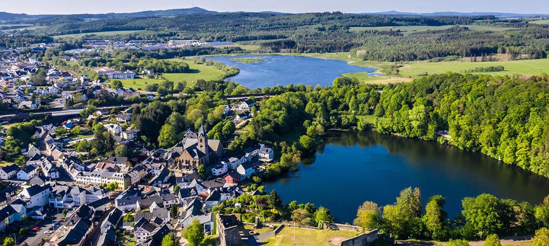 kurvenkreis-freizeit-ulmener-maar-und-jungerweiher-dominikketz_1080 Luftaufnahme einer Kleinstadt, umgeben von üppigem Grün und zwei Seen. Die Stadt zeichnet sich durch Häusergruppen mit weißen und grauen Dächern aus. Waldgebiete und sanfte Hügel erstrecken sich unter einem klaren blauen Himmel in die Ferne.