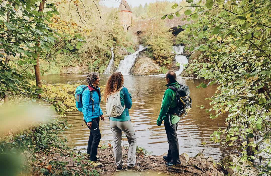 kurvenkreis-freizeit-wandern-schieferland-kaisersesch_1080 Drei Wanderer stehen am Flussufer und betrachten einen Wasserfall und ein kleines burgähnliches Bauwerk in einem üppigen, grünen Wald. Sie tragen Outdoor-Ausrüstung und Rucksäcke. Bäume rahmen die Szene ein.